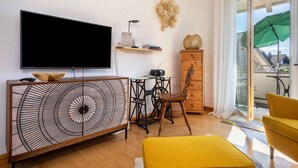 A living room with a flat-screen TV, a wooden cabinet with a geometric design, a small table with a lamp, a chair, and a yellow ottoman. - Apartment 'Haus Grete' with Lake View and Balcony (Langenargen)