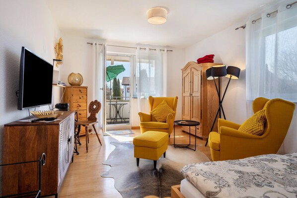 Habitación de hotel moderna con televisión de pantalla plana, muebles de madera, un sillón amarillo, una cama con estampado y vistas a través de una puerta de cristal.