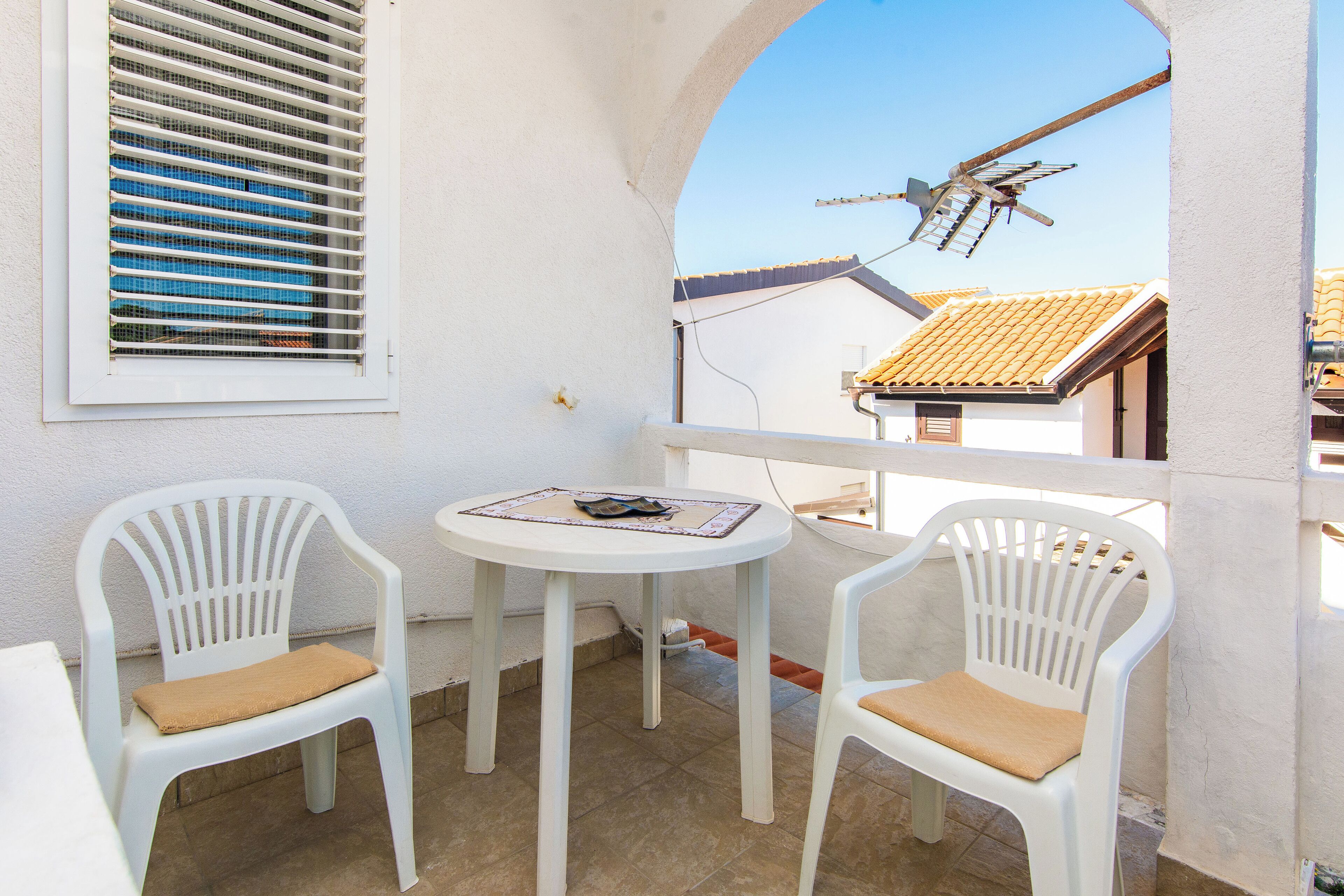 A balcony with two white plastic chairs and a small round table, a window with blinds, and a view of a building with a tiled roof.