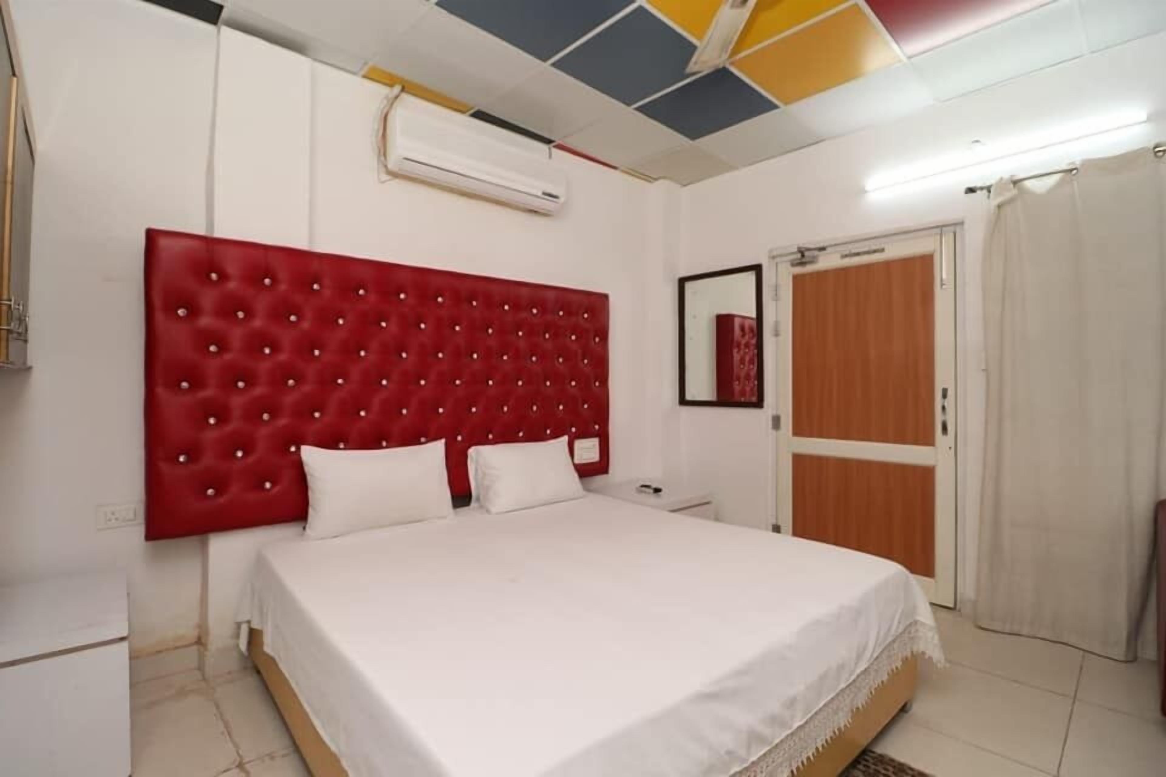 A bedroom with a red tufted headboard, white bedding, a wooden wardrobe, and a white air conditioning unit.