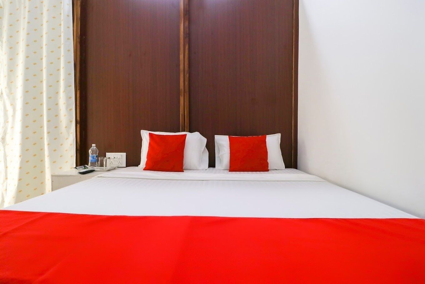 A hotel room with a bed, two red and white pillows, a wooden wardrobe, and a white curtain.