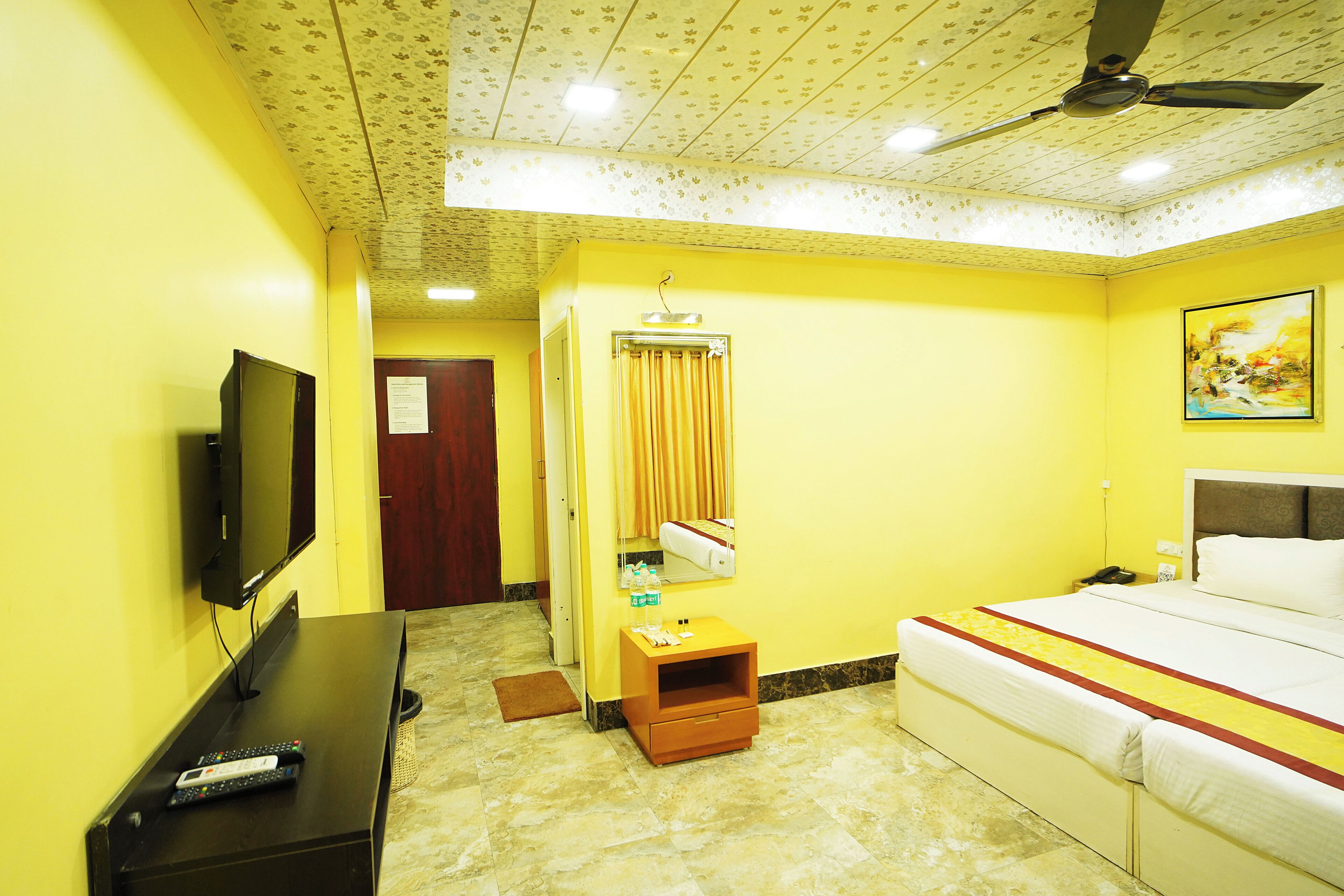 A hotel room with a bed, a television, a small table, a mirror, a ceiling fan, and a door.