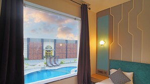 Habitación de hotel con cama, ventana con cortinas, vista a la piscina y una lámpara de pared.
