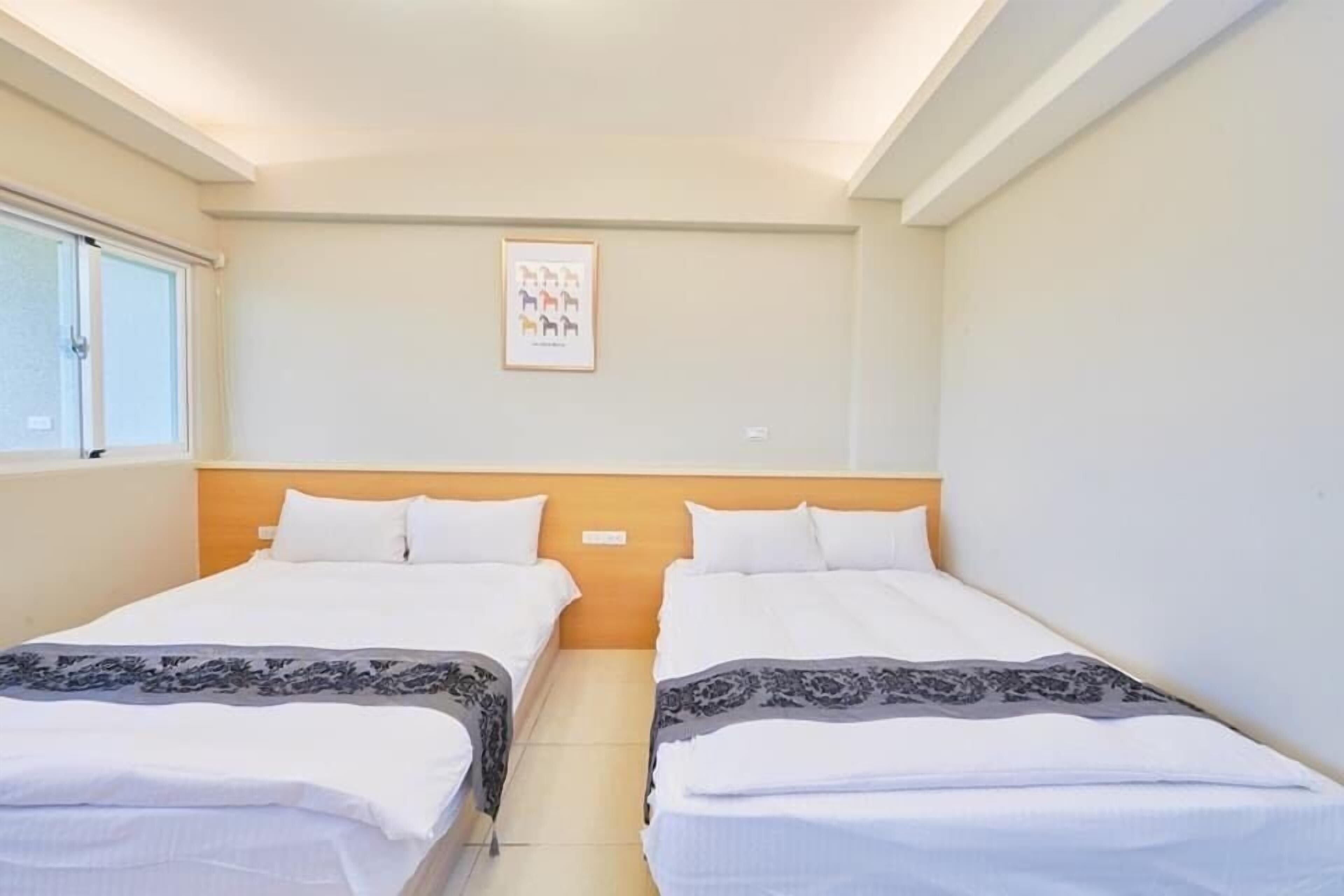 A hotel room with two single beds, a wooden headboard, a framed picture on the wall, and a window on the left.
