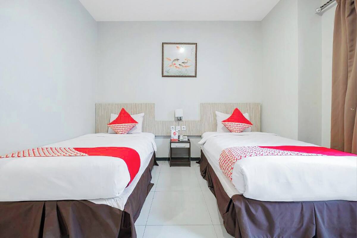 A hotel room with two beds, each with red and white bedding, a small bedside table with a lamp, and a framed artwork on the wall.