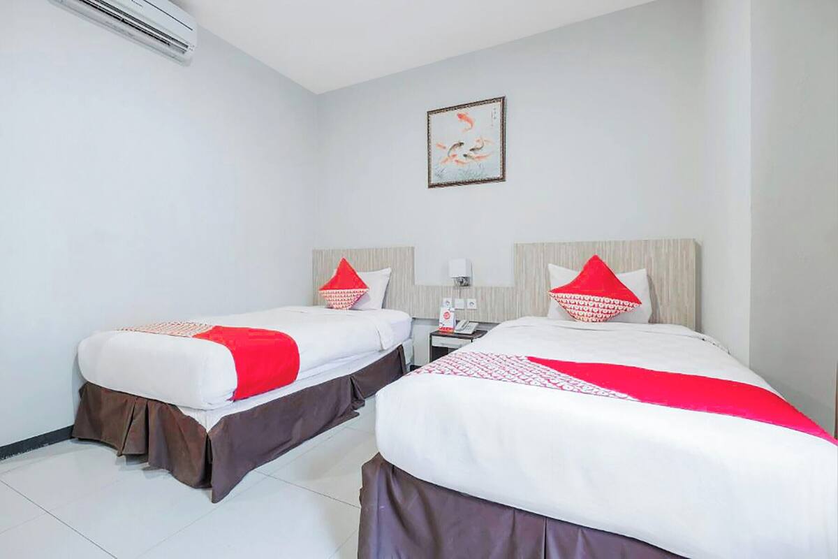 A hotel room with two beds, each with a red and white bedspread and matching pillows. There is a small bedside table with a lamp and a phone. The room has white walls and a framed picture on the wall.