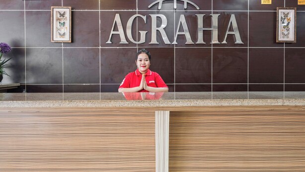 A reception desk with a woman in a red uniform, a large 'AGRAHA' sign, framed artwork, and clocks on the wall. - Hotel O Marina Andalas (Makassar)