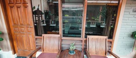 A wooden porch with two chairs and a table, a wooden door, and a window with a view of the garden.