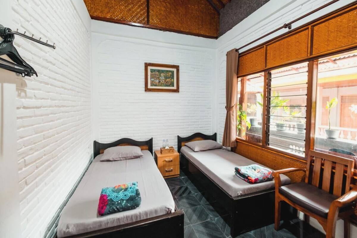 A small, cozy room with two single beds, a wooden chair, a nightstand, and a window with curtains.