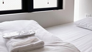 Una cama bien hecha con sábanas blancas, una toalla doblada y una bolsa de plástico sobre la cama. Se ve una ventana con marcos negros y vistas al exterior.