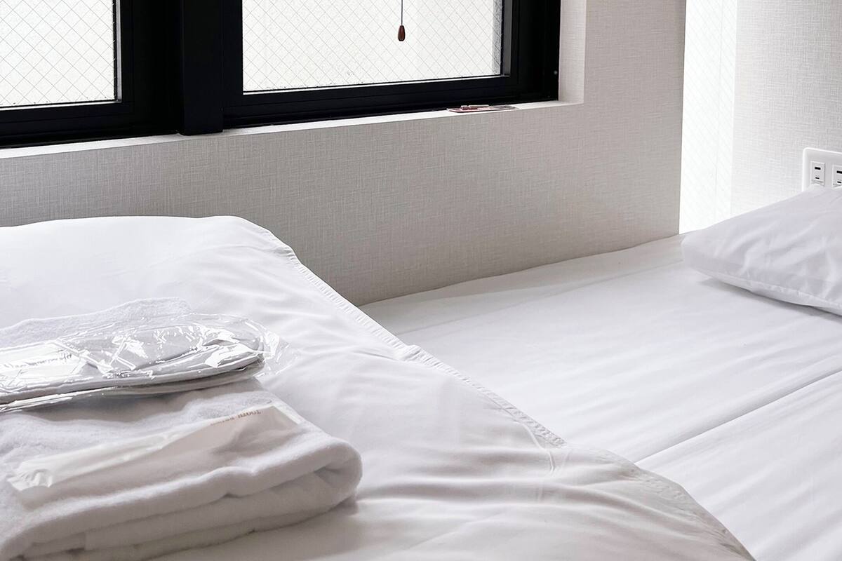 Una cama bien hecha con sábanas blancas, una toalla doblada y una bolsa de plástico sobre la cama. Se ve una ventana con marcos negros y vistas al exterior.