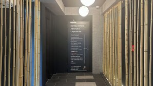 Entrada de hotel con un muro de bambú, un letrero que enumera servicios como gimnasio y baños públicos, y una escalera que conduce a la recepción.