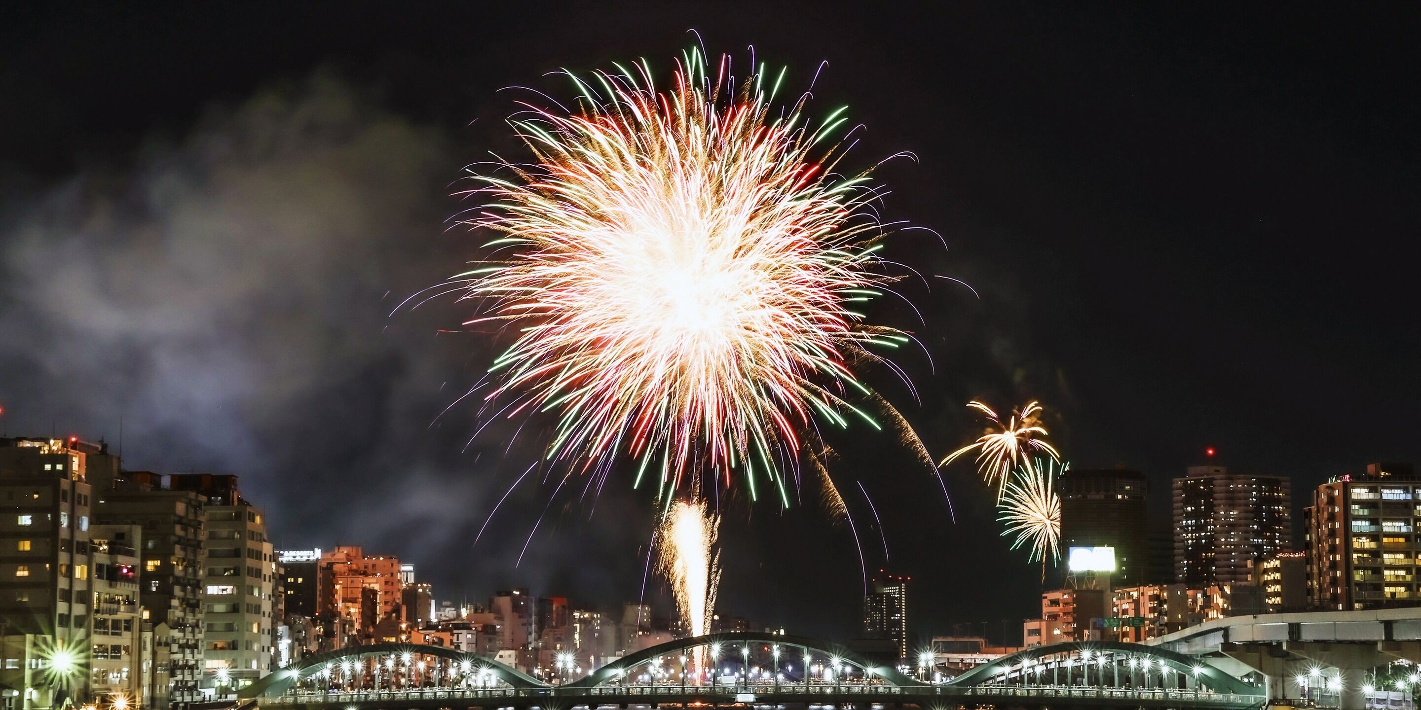 A nighttime cityscape with fireworks, a bridge, and illuminated buildings.