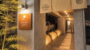 Entrada de un hotel con un cartel que indica Hotel Winds Asakusa, un albergue tipo cápsula, y un camino que conduce al interior.