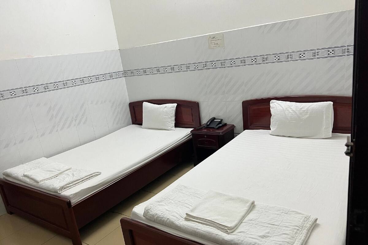Habitación de hotel con dos camas individuales, cada una con cabecero de madera y ropa blanca. Hay una mesita de noche pequeña con un teléfono y una lámpara. Las paredes están cubiertas de azulejos con un diseño geométrico, y hay un friso decorativo a lo largo del techo.