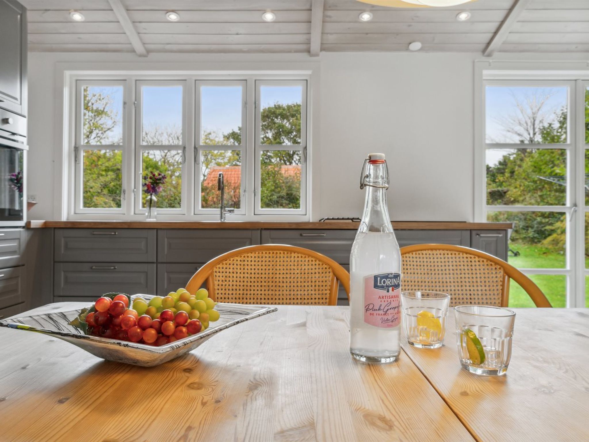 A kitchen with a wooden table, a bottle of liquor, glasses, and a bowl of fruit.