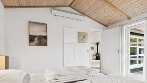Un dormitorio con techo de madera, paredes blancas y un ventanal grande. Hay una cama con ropa de cama blanca y un cuadro en la pared.