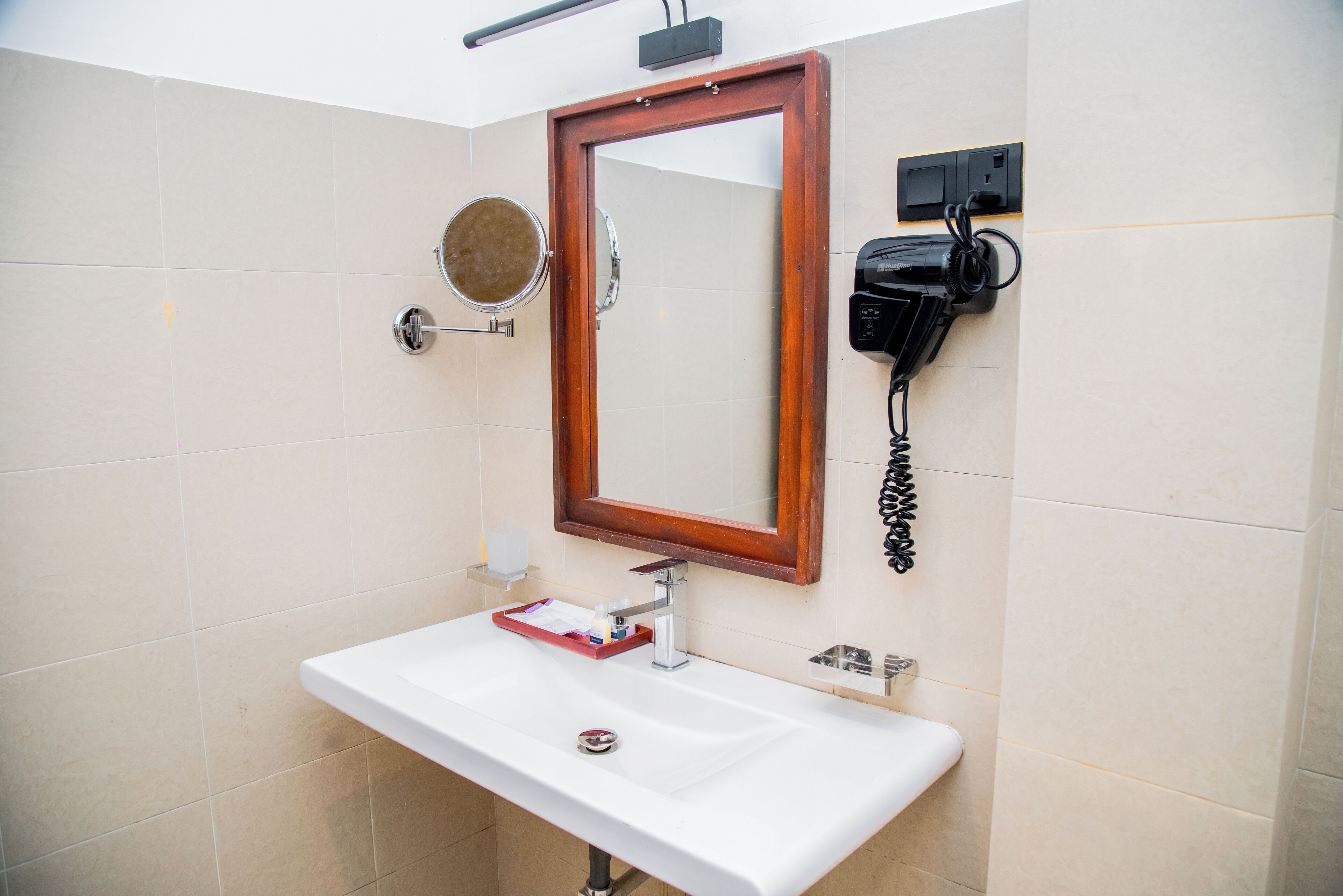 Een badkamer met een wit wastafelmeubel, een spiegel met houten lijst, een ronde spiegel en een haardroger bevestigd aan de muur.