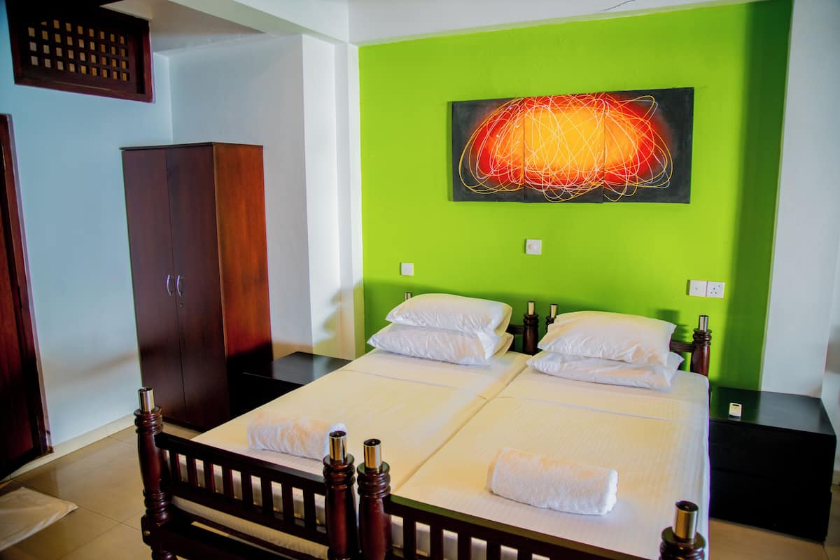 A double bed with white linens and two pillows, a wooden wardrobe, and a green accent wall with a painting.
