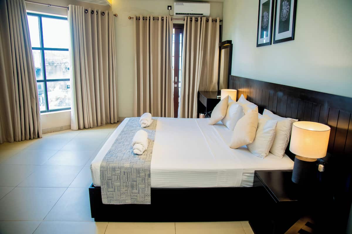 A hotel room with a large bed, a bedside table with a lamp, a desk with a chair, and a window with curtains.