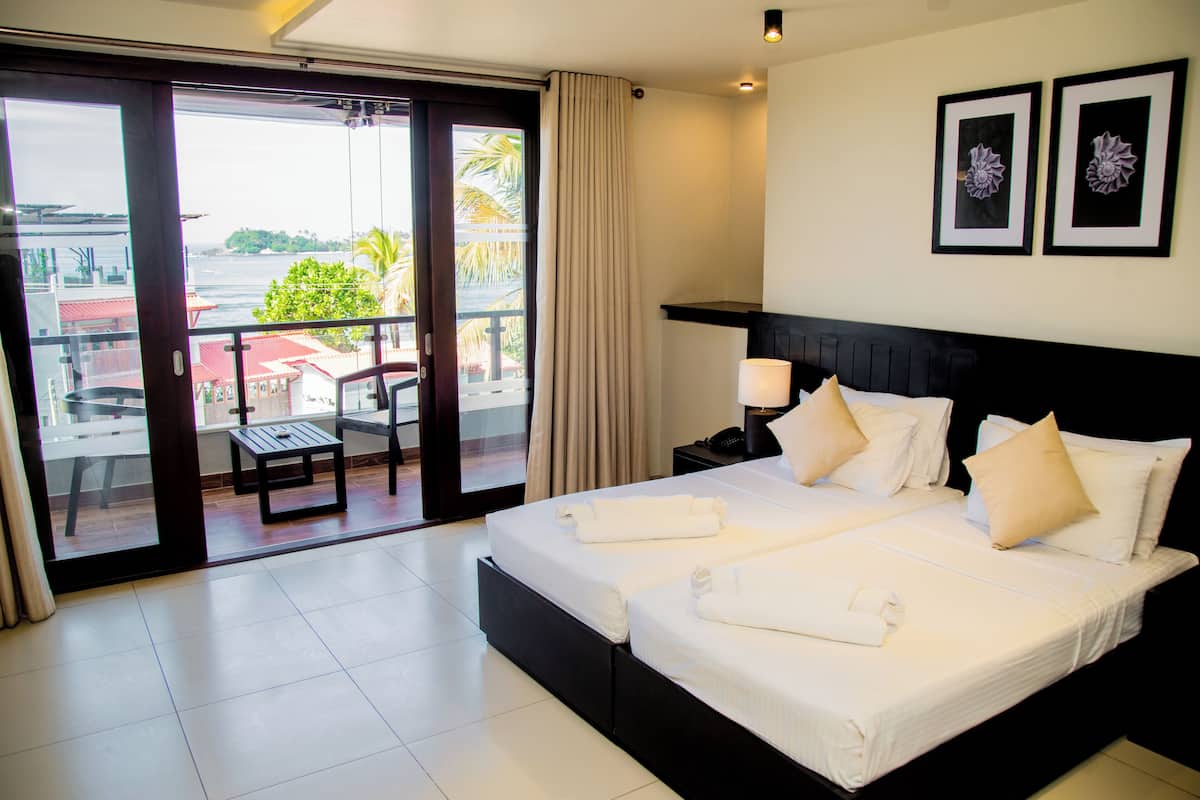 A hotel room with a large bed, two bedside tables, a desk, and a chair. There is a balcony with a view of the sea and palm trees.