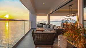 Un balcón con sofá, sillas y mesa, con vista a un cuerpo de agua y un paisaje urbano al atardecer.