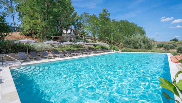 A rectangular swimming pool with clear blue water, surrounded by lounge chairs and umbrellas, set against a backdrop of trees and a rocky hillside. - Lovely apartment with pool, A/C, WIFI, TV, veranda and panoramic view, close to San Gimignano (Montaione)