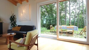 Una sala de estar con un sofá, una silla de madera, una mesita y un amplio ventanal corredera que da a un área de comedor exterior con mesa y sillas.