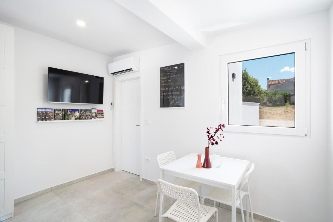 A modern, white-walled room with a flat-screen TV, a chalkboard with writing, a window with a view, and a dining table with chairs.