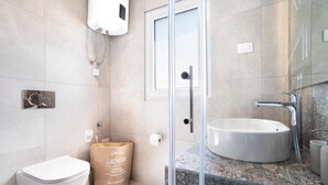 Un baño moderno con cabina de ducha de vidrio, inodoro blanco, una bolsa de lavandería grande color beige y un lavamanos redondo con encimera de granito.