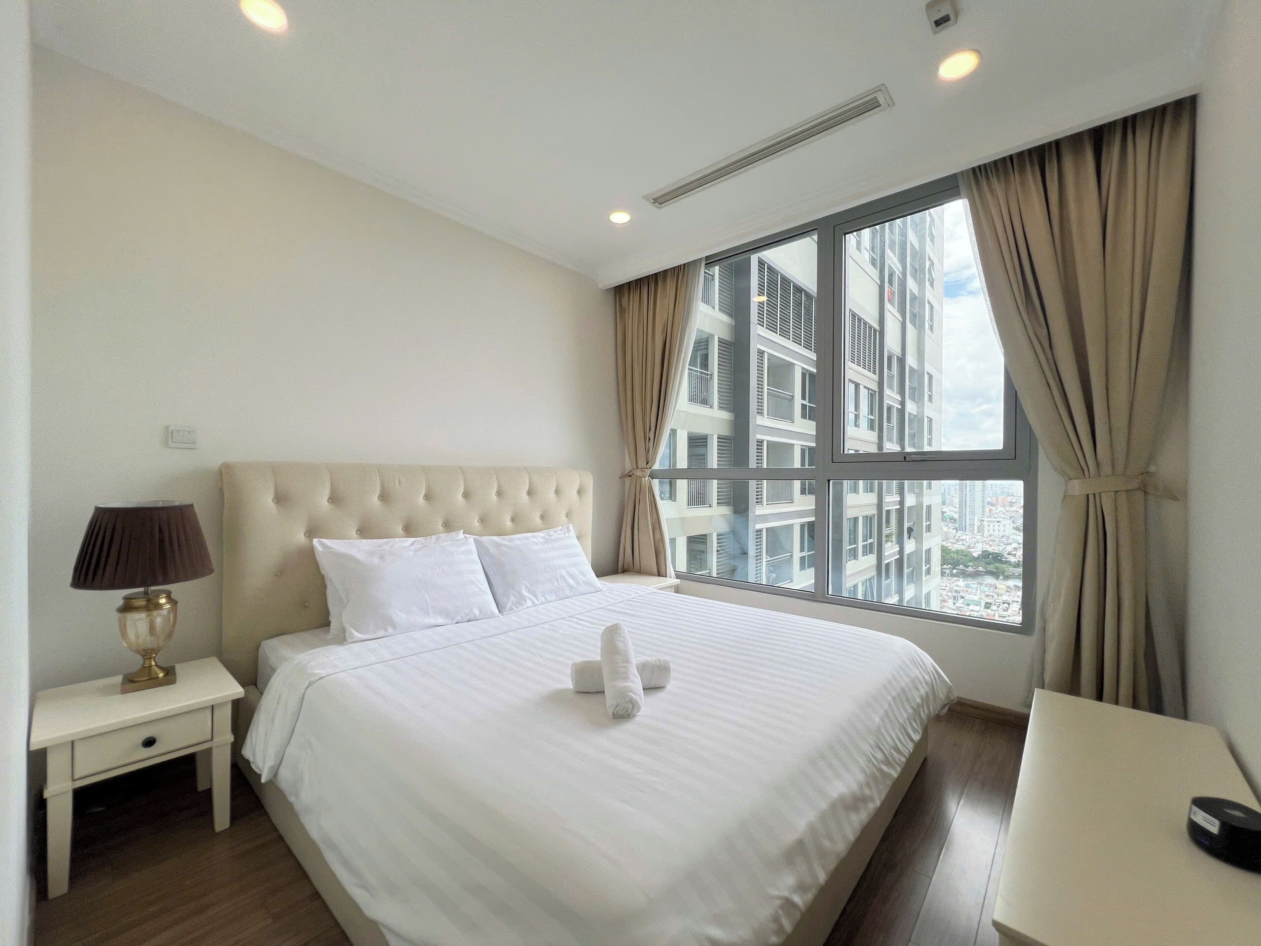 A hotel room with a large bed, a bedside table with a lamp, a window with curtains, and a view of buildings.