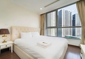 A hotel room with a large bed, a nightstand with a lamp, and a view of tall buildings through a large window.