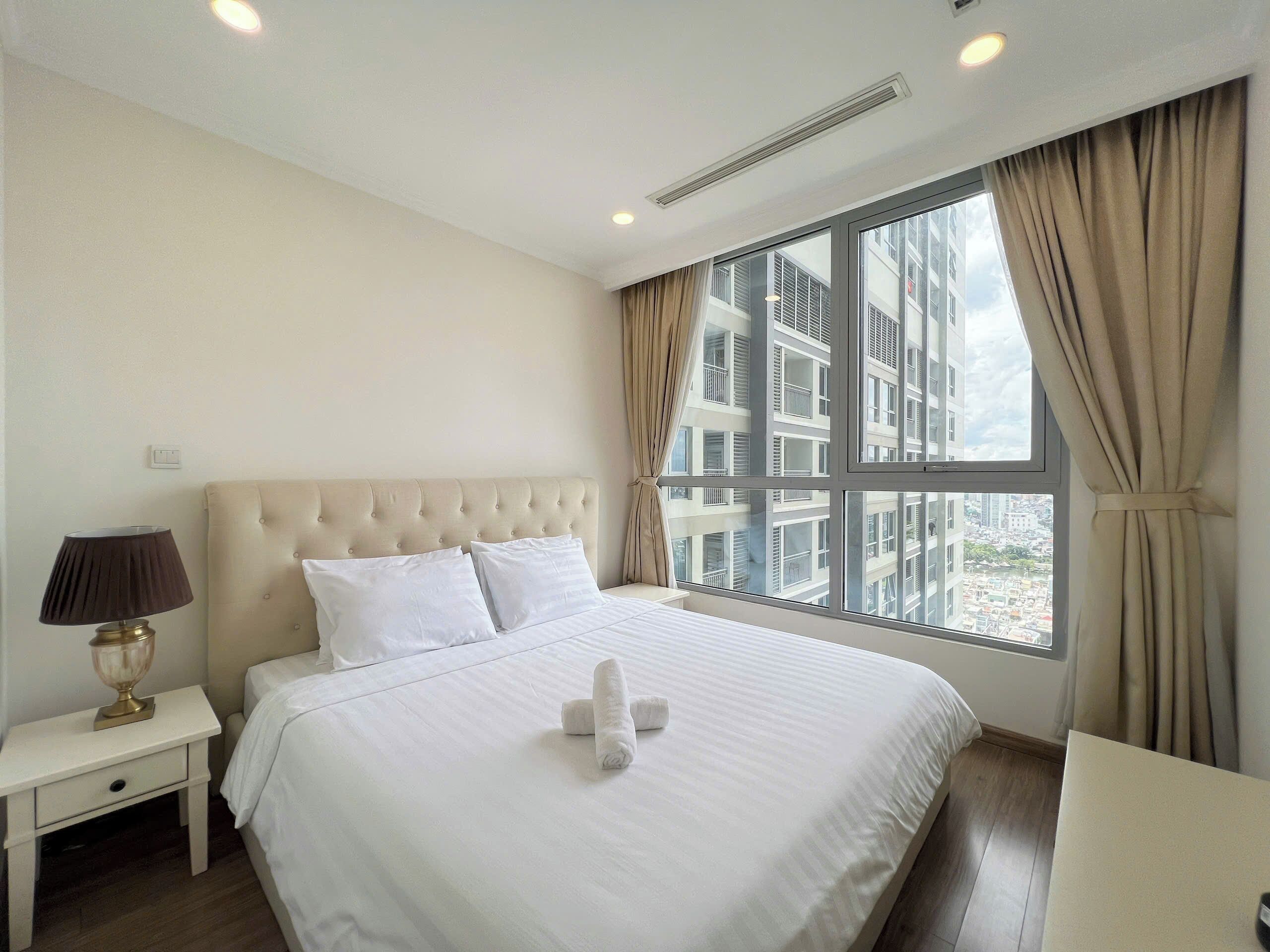 A hotel room with a large bed, a nightstand, a lamp, and a view of a building through the window.
