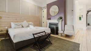 Una camera da letto con una testiera in legno, un letto grande, uno specchio rotondo, un caminetto e un angolo salotto con due poltrone.