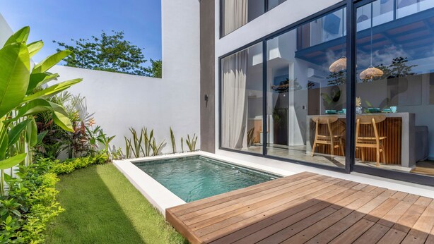 A modern outdoor pool area with a wooden deck, surrounded by lush greenery and a glass-enclosed patio. - Dela Villa by Betterplace (Pecatu)