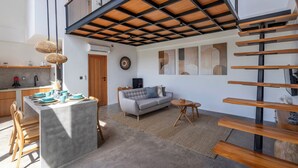 A modern kitchen with a central island, a dining area with a sofa, and a staircase with wooden beams. - Dela Villa by Betterplace (Pecatu)
