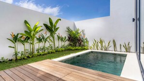 A modern outdoor pool area with a wooden deck, surrounded by lush greenery and a white wall. - Dela Villa by Betterplace (Pecatu)