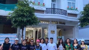 Un groupe de personnes se tient devant un immeuble doté d’un balcon et d’une enseigne sur laquelle on peut lire « HÔTEL ».