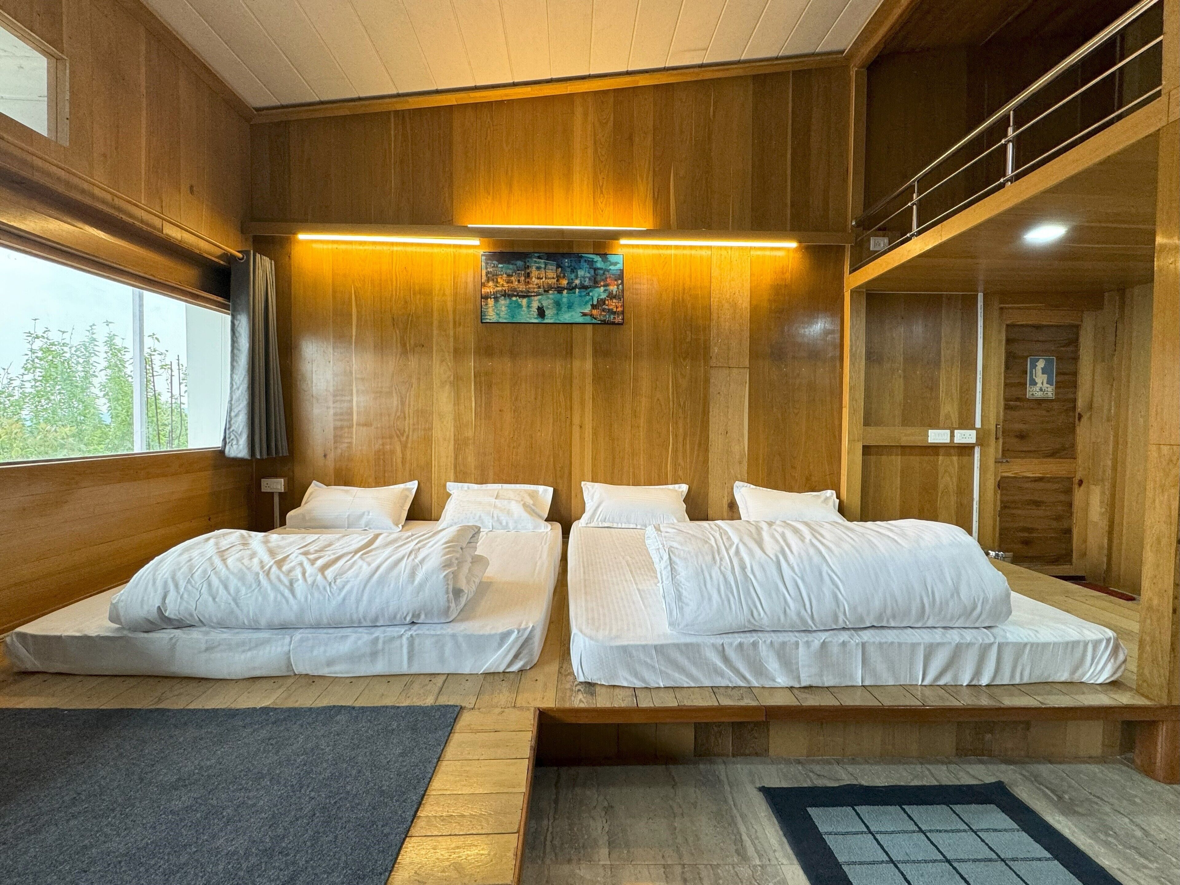 Una habitación con dos camas, paredes de madera, un cuadro sobre las camas y una ventana con vistas a árboles.