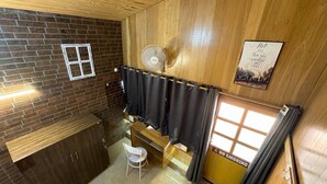 A small room with a wooden ceiling, a brick wall, a window with curtains, a fan, a small table, and a chair. - Wanderlust Mukteshwar (Dhari)
