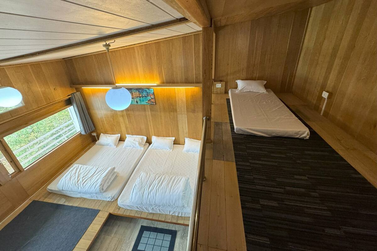 Une pièce aux murs et au plancher en bois, avec deux lits recouverts de draps blancs, une petite table et une fenêtre donnant sur un paysage verdoyant.