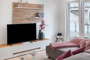 Una sala de estar moderna con un televisor de pantalla plana, estanterías de madera, un florero con flores y un sofá con una manta rosa.