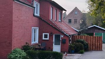 Ein rotes Backhaus mit geneigtem Dach, einer Haustür und einer kleinen Veranda mit Tisch und Stuhl.