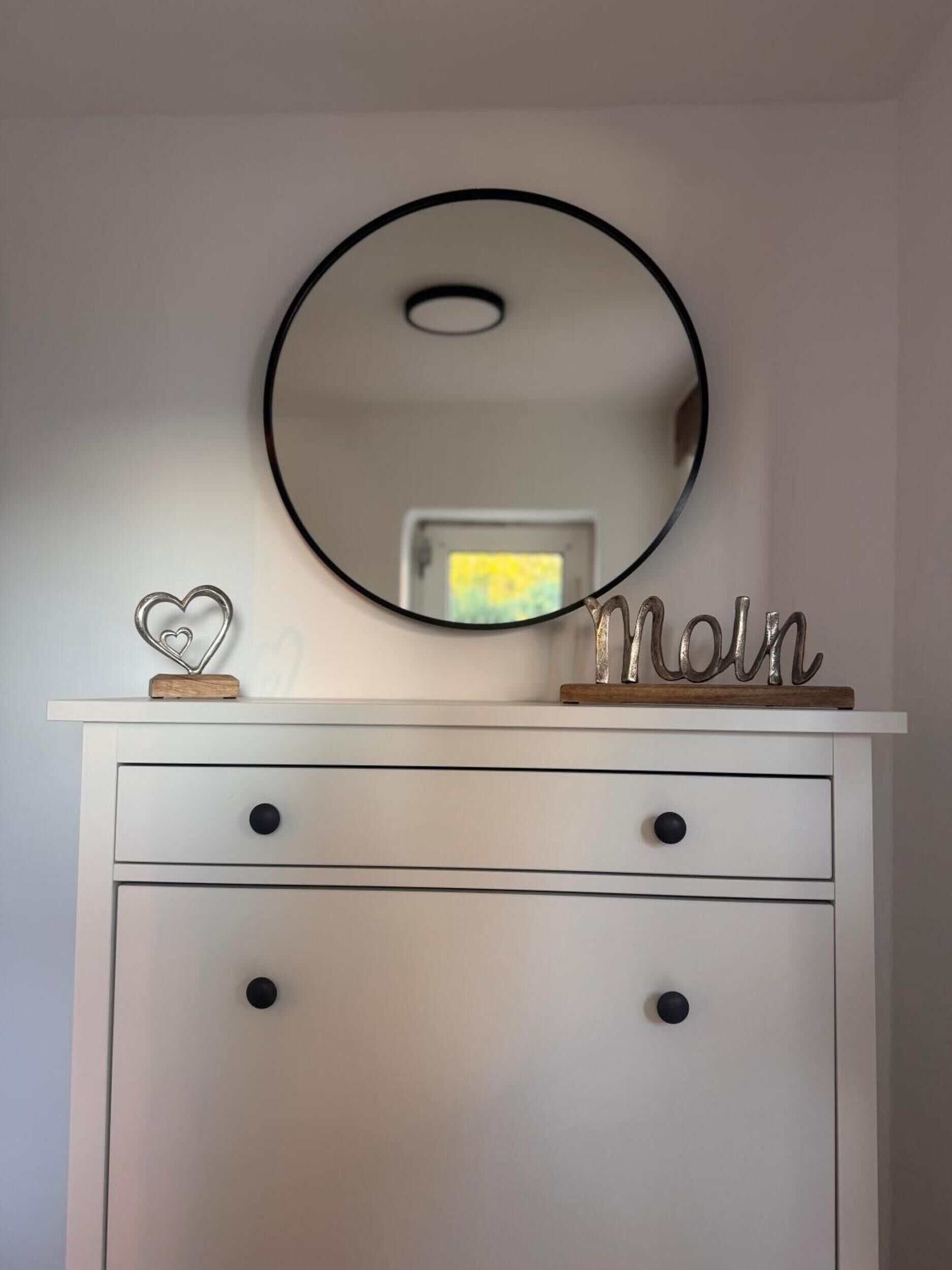 Un aparador blanco con un espejo redondo encima, una decoración con forma de corazón y un cartel de metal con la palabra 'Molin'.