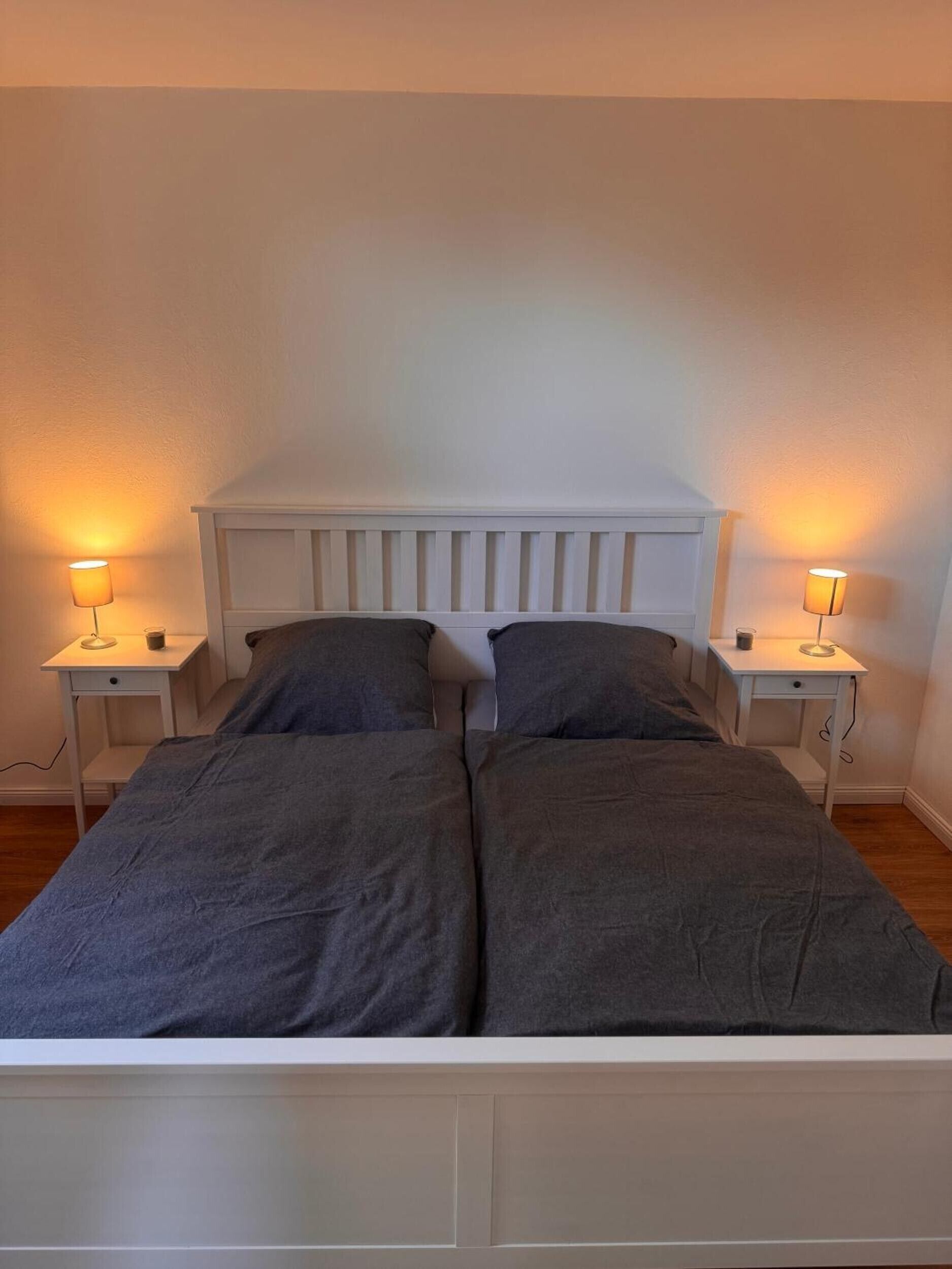 A double bed with a white headboard, two bedside tables with lamps, and a plain wall.