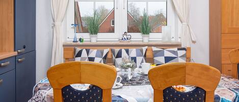 Un comedor con una mesa lista para dos personas, una ventana con cortinas y vistas a casas.