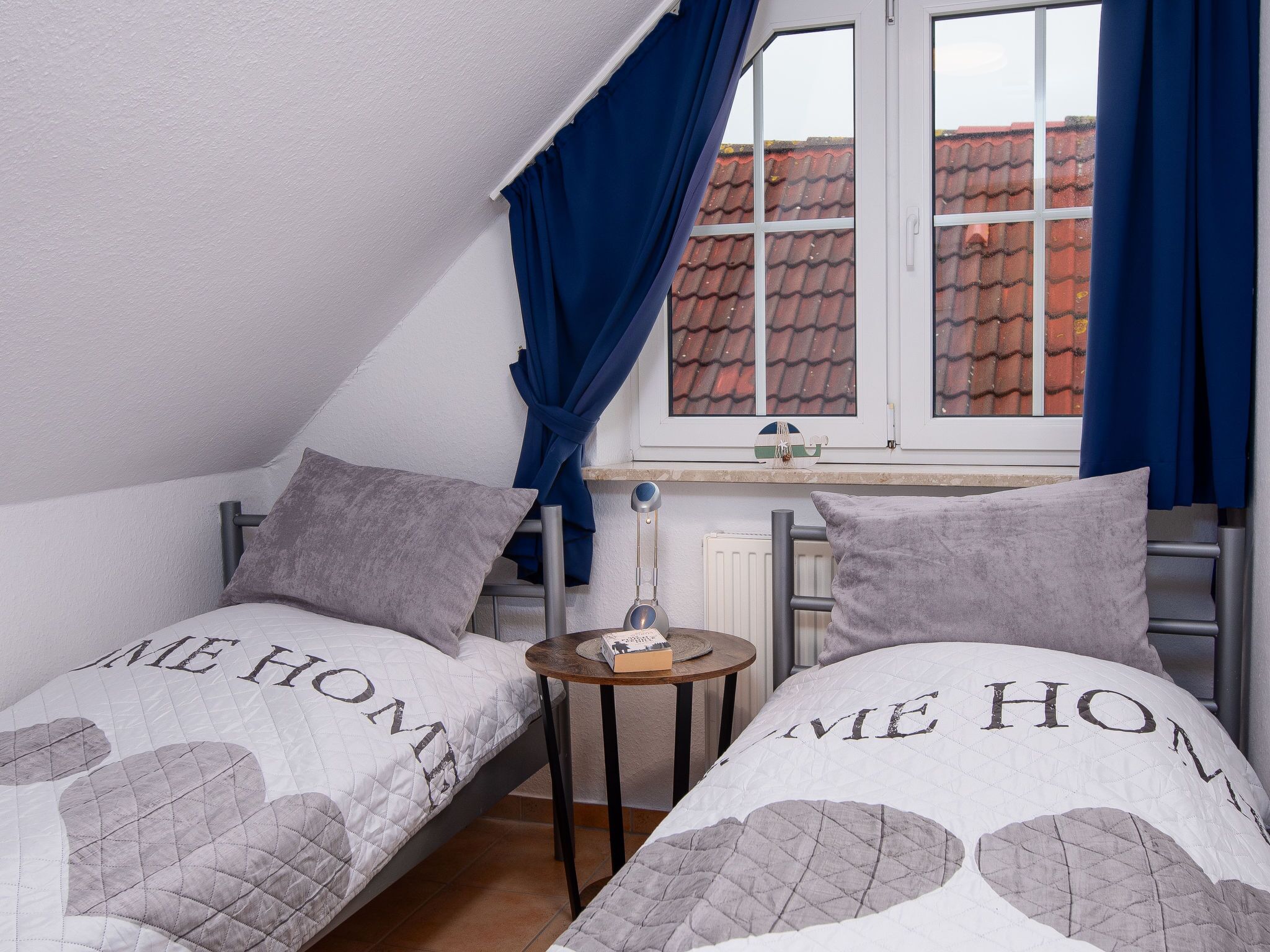 A small, cozy room with two beds, each with a quilted blanket featuring the words "HOME". There is a window with blue curtains, a small table with a lamp, and a radiator.