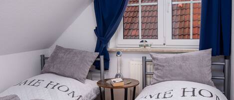 A small, cozy room with two beds, each with a quilted blanket featuring the words "HOME". There is a window with blue curtains, a small table with a lamp, and a radiator.