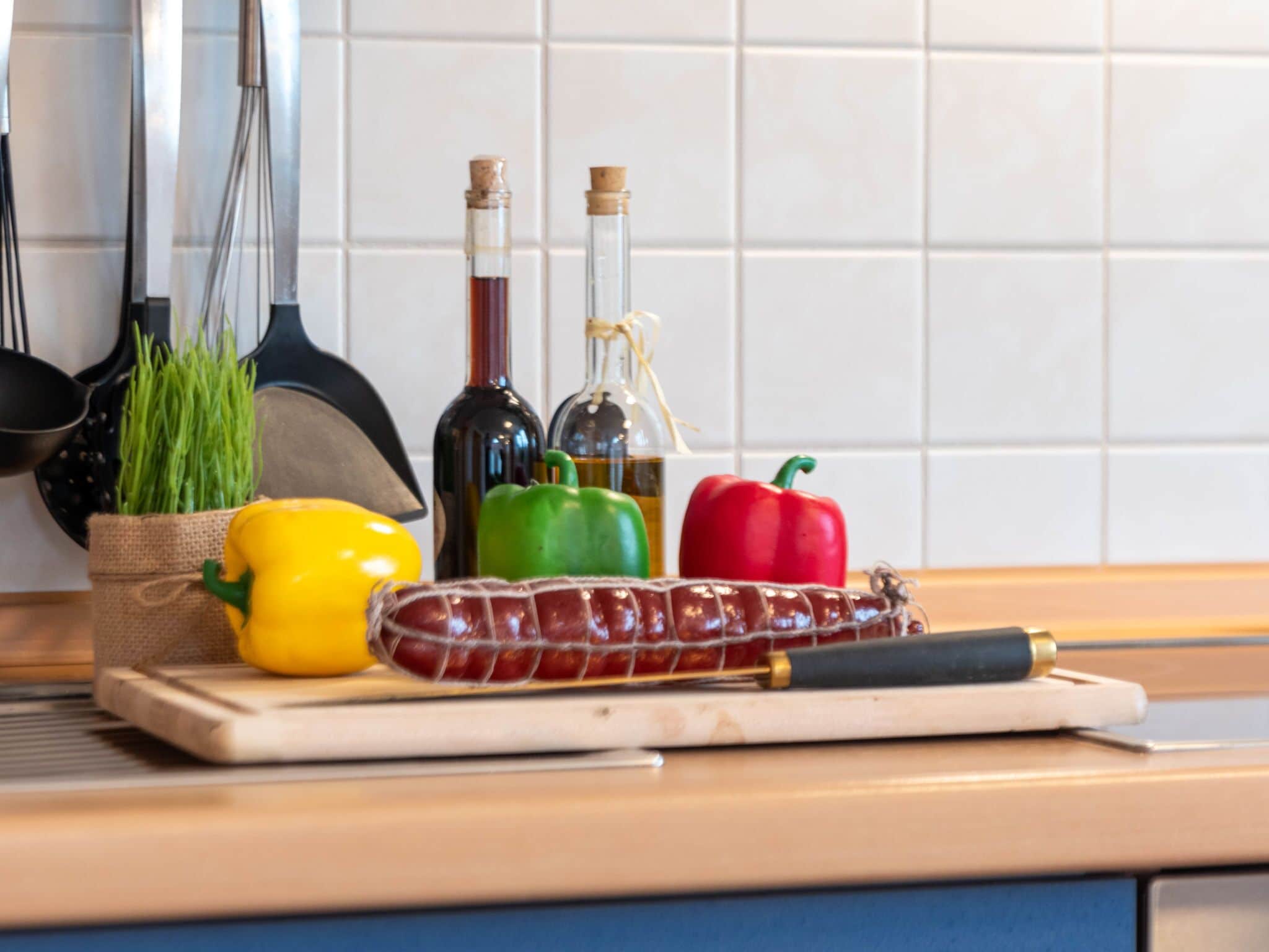 Eine Küchenarbeitsplatte mit Schneidebrett, einer gelben Paprika, einer grünen Paprika, einer roten Paprika, einer Wurst und zwei Flaschen Olivenöl.