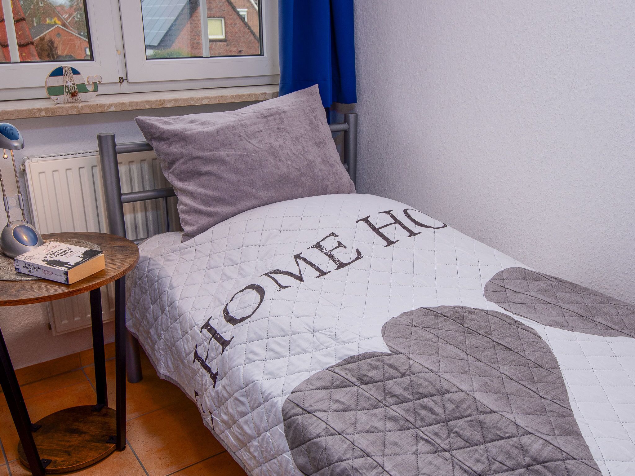 Cama bem arrumada com manta acolchoada e travesseiro, mesa de cabeceira com abajur e um livro, e uma janela com cortinas.